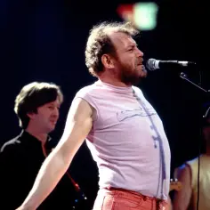 Joe Cocker śpiewa na scenie, wykonując swoje legendarne utwory. W tle widać innych muzyków.