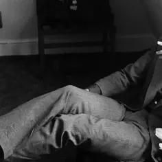 Uśmiechnięty Leonard Cohen, palący papierosa, w garniturze. W tle kwieciste tkaniny, jakby inspirowane jego ponadczasowymi utworami.