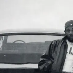 Kendrick Lamar, w czapce i skórzanej kurtce, opiera się o samochód. Jego styl przypomina okładki jego albumów.