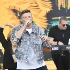 Wokalista na scenie, wykonujący utwory. W tle klawiszowiec i tancerka.