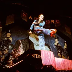 Elton John w błyszczącym stroju, kopiący nogą na scenie, wykonuje swoje największe przeboje.