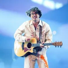 Artysta w okularach gra na gitarze, tworząc fancy utwory. Jego ubranie ozdobione jest pomarańczowymi płomieniami.