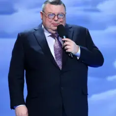 Mężczyzna w garniturze i okularach przemawia na scenie podczas turnieju piosenki.