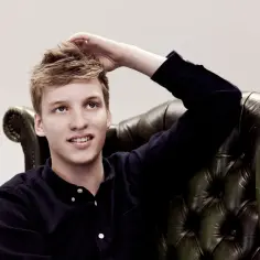 George Ezra w fotelu, z ręką na głowie, jakby zastanawiał się nad tekstem piosenki "Shotgun".