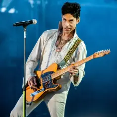 Prince gra na gitarze, wykonując swoje legendarne utwory. Wokalista w białej, zdobionej koszuli i spodniach, z charakterystyczną fryzurą, skupiony na muzyce.
