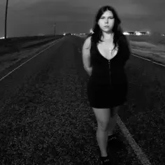 Dziewczyna w czarnej sukience stoi na pustej drodze nocą. Może to scena z teledysku "Sailor Song" Gigi Perez.