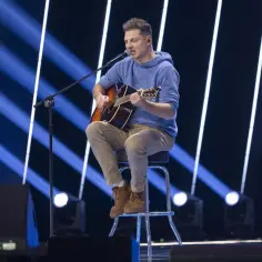 Mężczyzna siedzi na stołku, gra na gitarze i śpiewa. W tle niebieskie światła. Może to być występ z utworami o tematyce letni, chamski podryw.