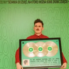 Dawid Podsiadło z potrójną platynową płytą za album "Małomiasteczkowy". Zielona ścianka z napisem "Małomiasteczkowa" stanowi tło.