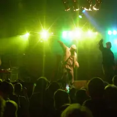 Koncert hip-hopowy. Na scenie raperzy, jeden z nich w czapce, jakby policjant. DJ przy stanowisku.