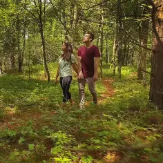 Para spaceruje w zielonym gaju, trzymając się za ręce. Słońce przebija się przez liście, tworząc magiczną atmosferę.
