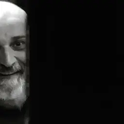 Uśmiechnięty Wojciech Sęp Dudziński, z charakterystyczną brodą i łysą głową, wychyla się zza ciemnej zasłony.