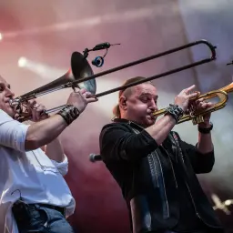 Dwóch muzyków Golec uOrkiestra gra na instrumentach dętych. W tle koncertowe światła.