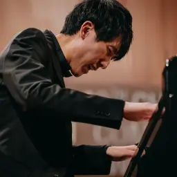 Pianista w czarnym garniturze gra na fortepianie, skupiony na wykonaniu utworu. Może to być jego występ podczas pierwszego konkursu chopinowskiego.