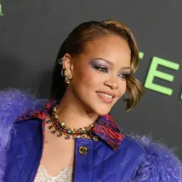 Rihanna w fioletowym futrze i błyszczącej biżuterii, jej wiek nie jest widoczny, ale stylizacja jest efektowna.