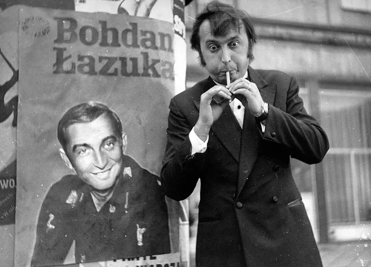 Bohdan Łazuka w garniturze, obok plakat z jego podobizną. Aktor z papierosem w ustach, wesoło pozuje do zdjęć.