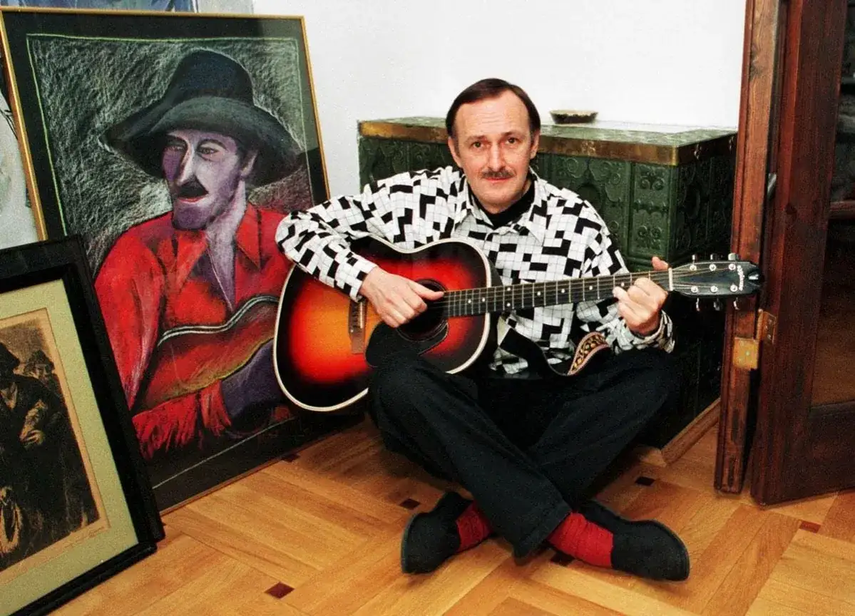 Jacek Cygan gra na gitarze przy portrecie swojego autorstwa. W tle widać fragmenty jego twórczości.