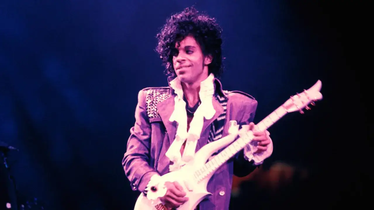 Prince gra na gitarze, wykonując swoje legendarne utwory. Jego charakterystyczna fryzura i fioletowa kurtka przyciągają wzrok.