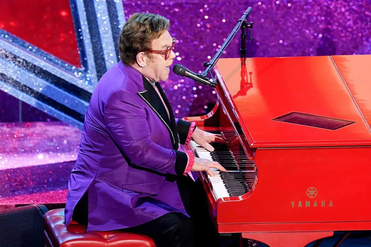 Elton John w fioletowej marynarce gra na czerwonym fortepianie, wykonując swoje największe przeboje.