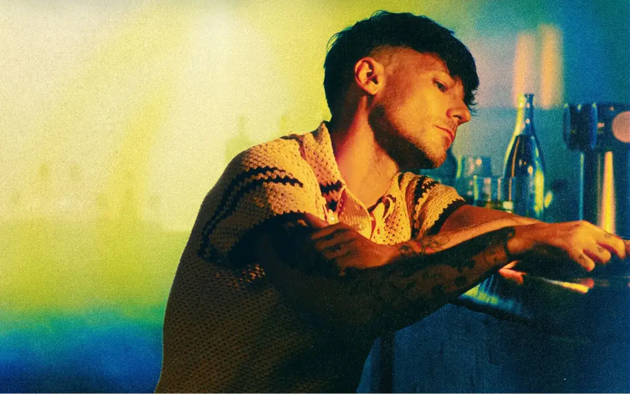 Louis Tomlinson w żółtym swetrze, z tatuażami na ramionach, przy barze. Jego utwory niosą emocje.