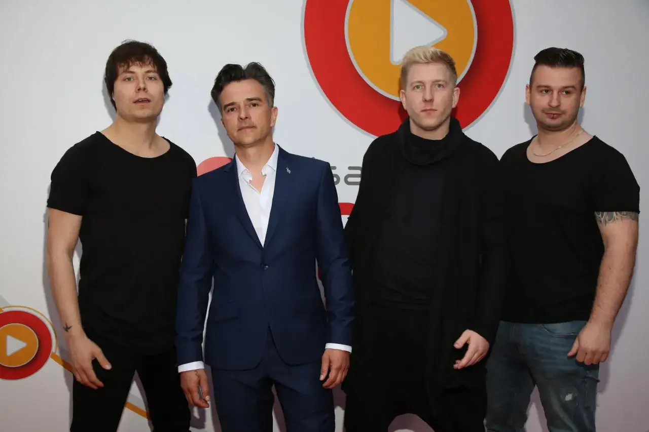 Czterech mężczyzn pozuje do zdjęcia. Dwóch w czarnych t-shirtach, jeden w granatowym garniturze, a czwarty w czarnej bluzie z kapturem. Wszyscy prezentują swoje loka utwory.