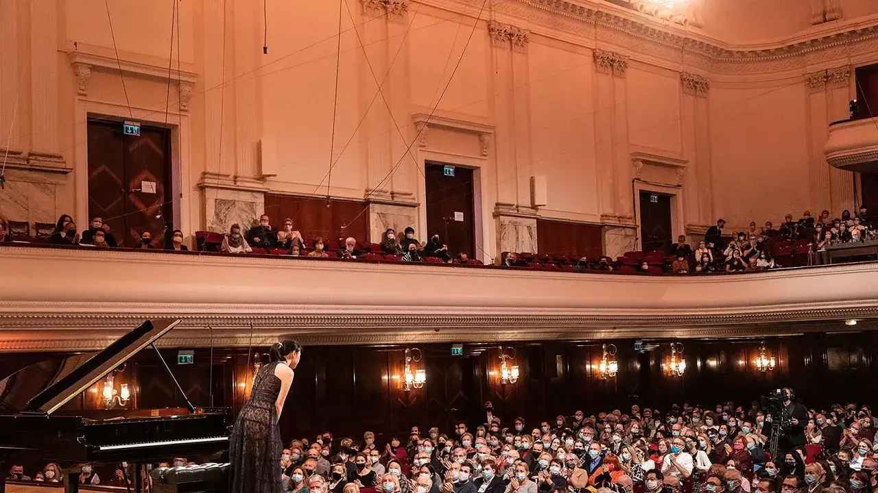 Widownia w operze podczas konkursu chopinowskiego. Pianistka w czarnej sukni przed fortepianem, publiczność w maskach na widowni i lożach.