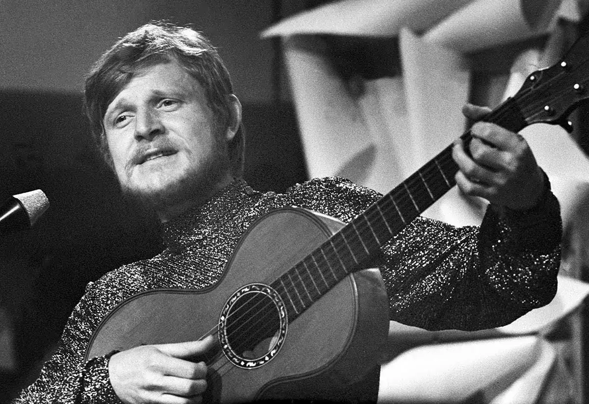 Piotr Szczepanik gra na gitarze, śpiewając swoje piosenki w błyszczącej marynarce.