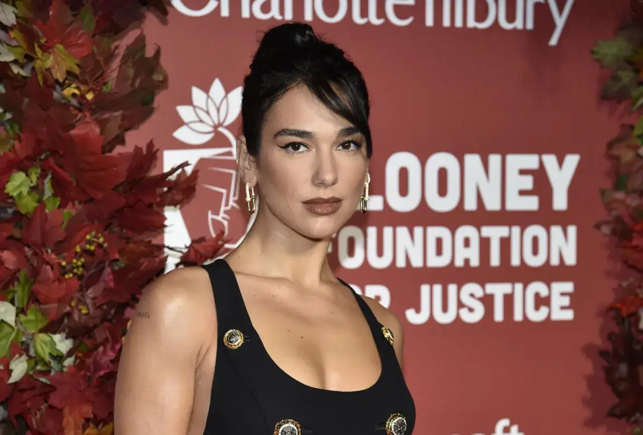 Dua Lipa, której pochodzenie jest albańskie, prezentuje się w czarnej sukience na tle czerwonej ściany z napisem 