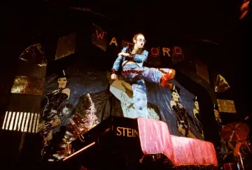 Elton John w błyszczącym stroju, kopiący nogą na scenie, wykonuje swoje największe przeboje.