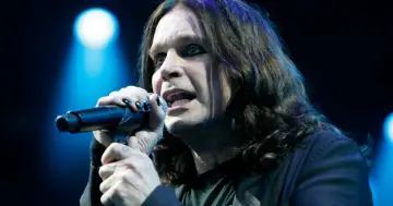 Ozzy Osbourne, mimo swojego wieku, wciąż śpiewa z pasją do mikrofonu na tle niebieskich świateł.