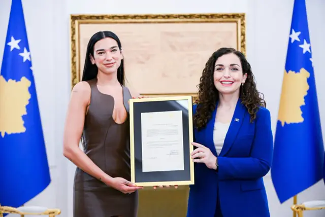 Dua Lipa, której dua lipa pochodzenie jest z Kosowa, odbiera dokument. Obok niej stoi kobieta w niebieskiej marynarce.
