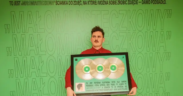 Dawid Podsiadło z potrójną platynową płytą za album "Małomiasteczkowy". Zielona ścianka z napisem "Małomiasteczkowa" stanowi tło.