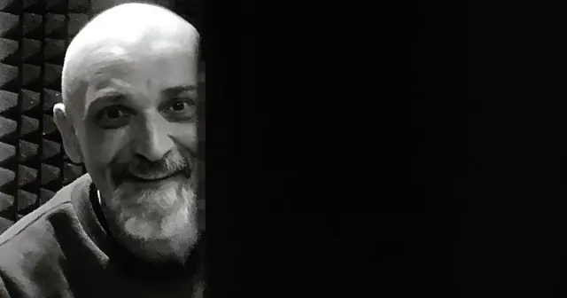 Uśmiechnięty Wojciech Sęp Dudziński, z charakterystyczną brodą i łysą głową, wychyla się zza ciemnej zasłony.